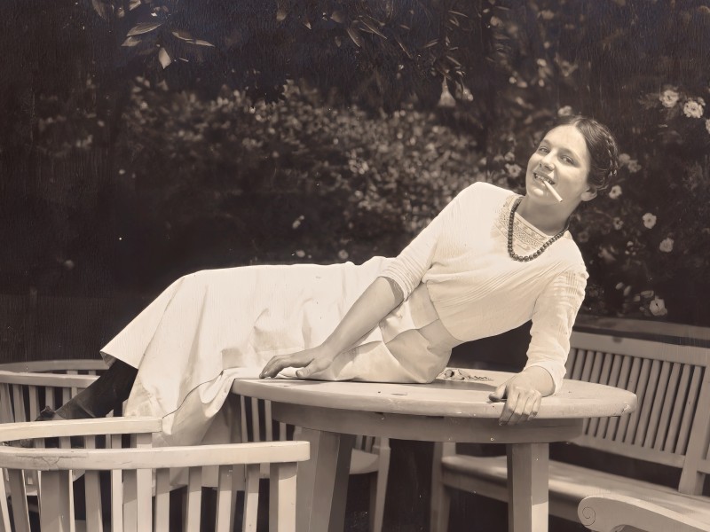 Frau in langem Kleid und Perlenkette liegt auf einem runden Holztisch, umgeben von Holzstühlen und einer Holzbank, im Garten mit Blumen und Büschen.