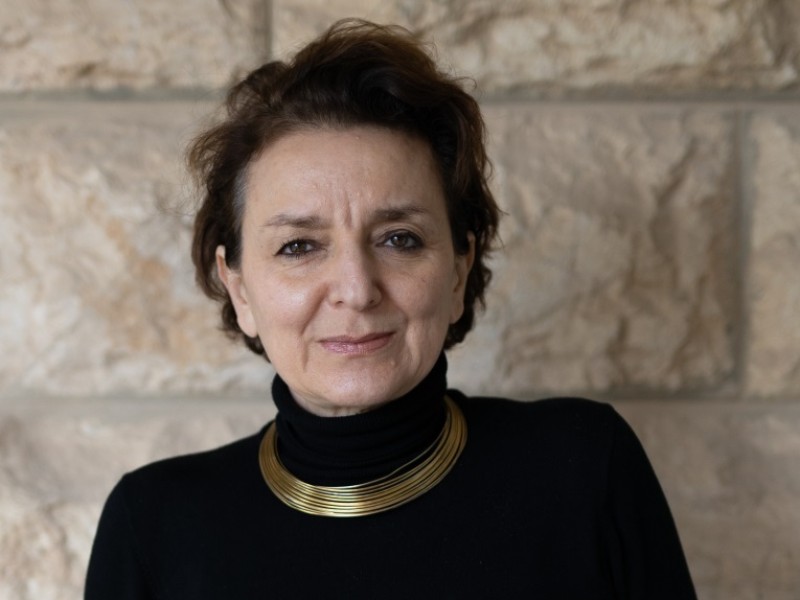 Woman standing in front of a stone wall, wearing a black turtleneck and a gold necklace, looking directly at the camera