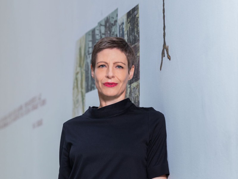 Barbara Staudinger standing against a wall with a metal menorah sculpture and a framed artwork in the background, wearing a black short-sleeve top and pants.