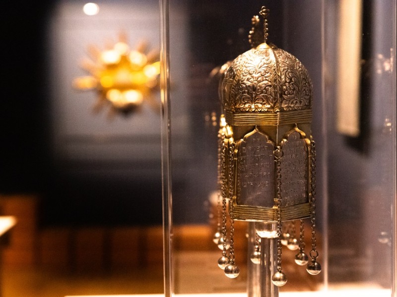 Ornate silver Torah finial with Hebrew inscriptions and hanging bells displayed in a museum case