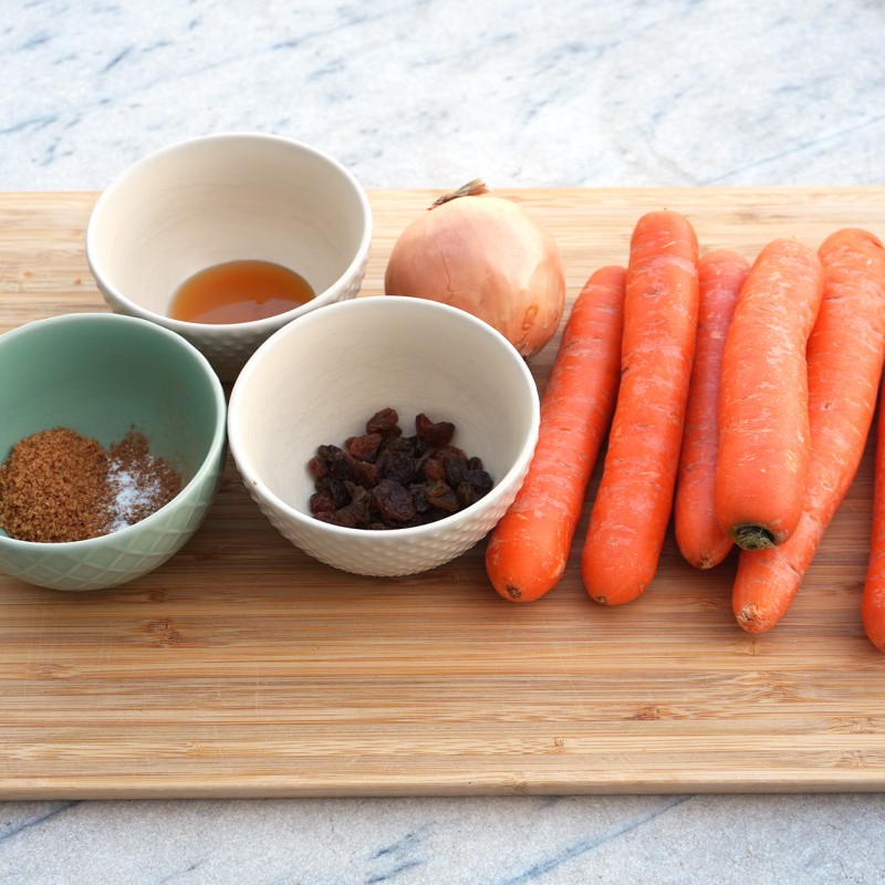 : Auf einem Holzbrett liegen sieben orangefarbene Karotten, eine Zwiebel, zwei Schalen mit Rosinen und einer braunen Zuckermischung sowie ein Glas mit einer Flüssigkeit.