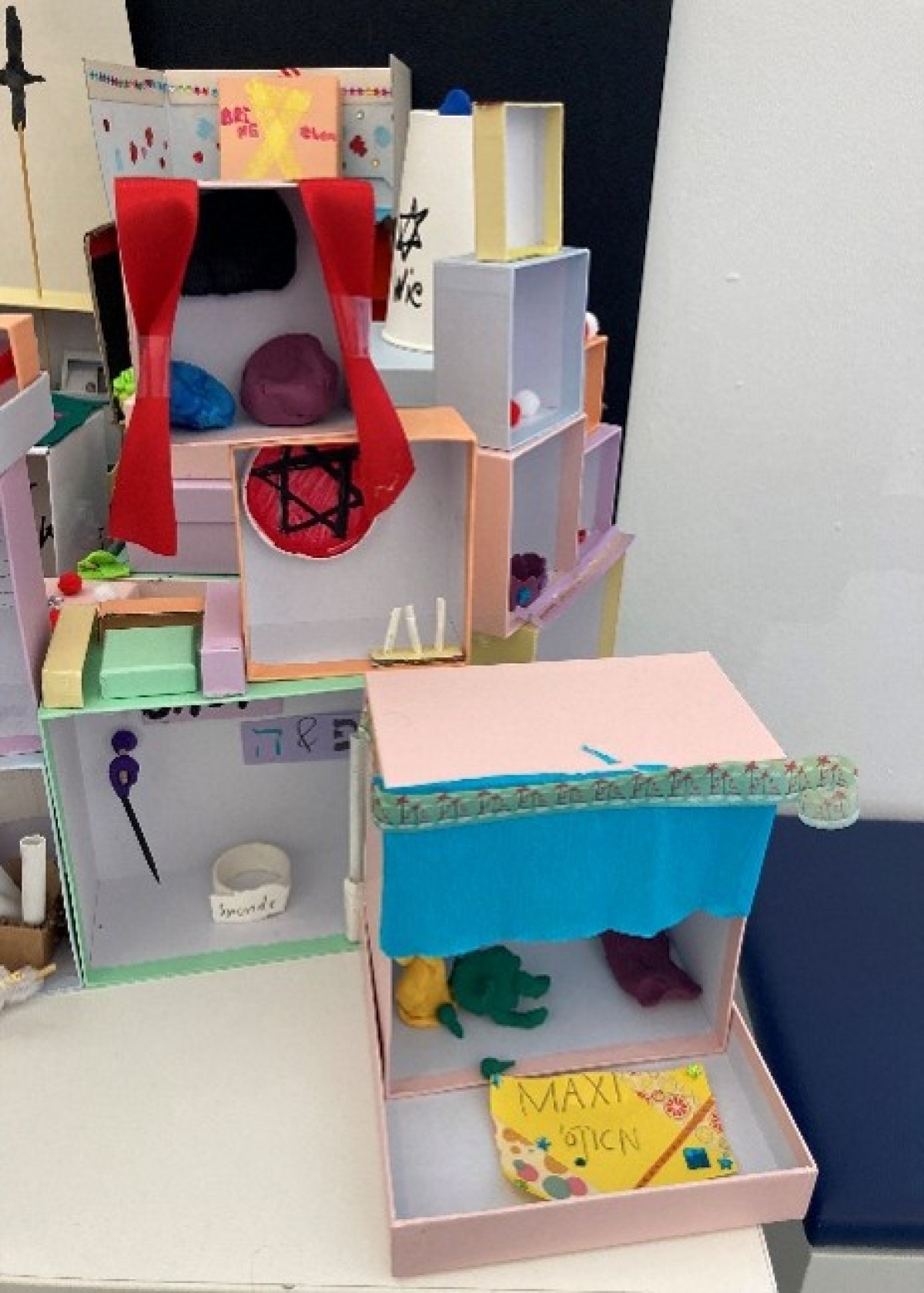 : Modell eines mehrstöckigen Puppenhauses aus Pappe mit bunten Details, Vorhängen und kleinen Figuren.