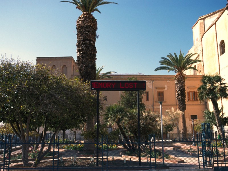 Städtischer Platz mit Palmen, Bäumen und einem digitalen Schild mit roter Schrift 'MEMORY LOST' vor einem historischen Gebäude.