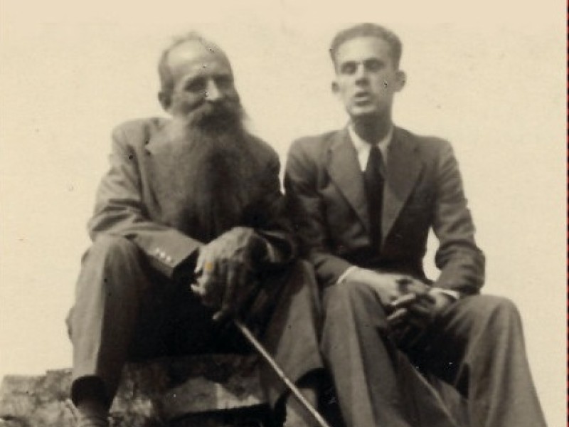 Zwei sitzende Personen in Anzügen auf einem Felsen, darunter rotes Buchcover mit Titel 'Du musst es für mich tun' von Eva Brenner.