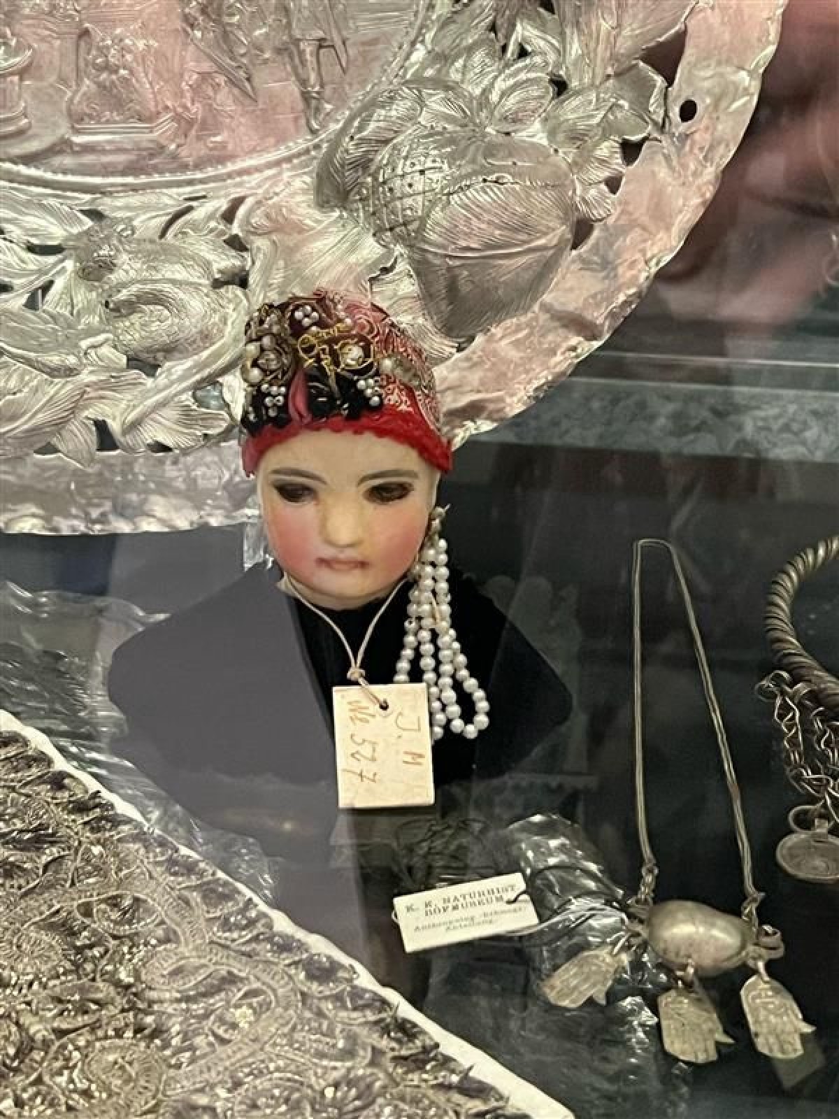 Traditionelle Figur mit Schmuck: Büste einer Figur mit roter, mit Perlen und goldfarbenen Schmuck verzierten Kopfbedeckung, umgeben von silbernen Schmuckstücken und einer reich verzierten silbernen Schale