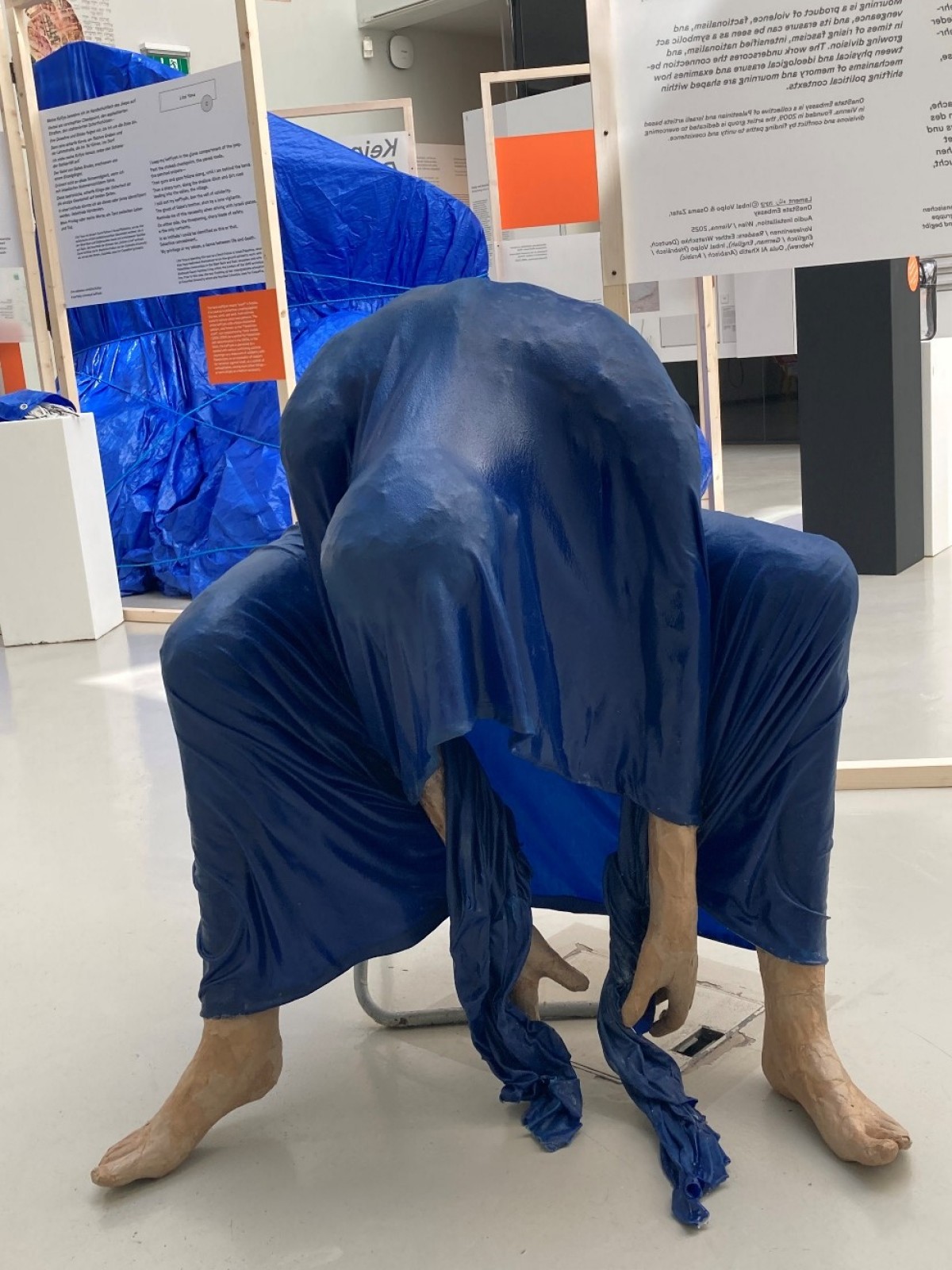 : Eine Skulptur einer weiblichen Figur, die unter einem blauen Schleier sitzt und sich nach vorne beugt.