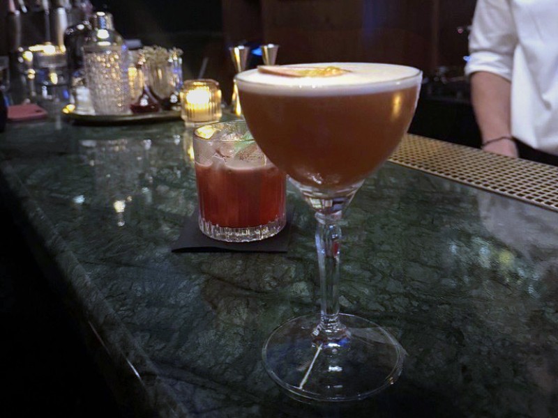 Zwei Cocktails auf einer dunklen Bar, einer in einem hohen Glas mit Schaum, der andere in einem kurzen Glas mit Eiswürfeln.