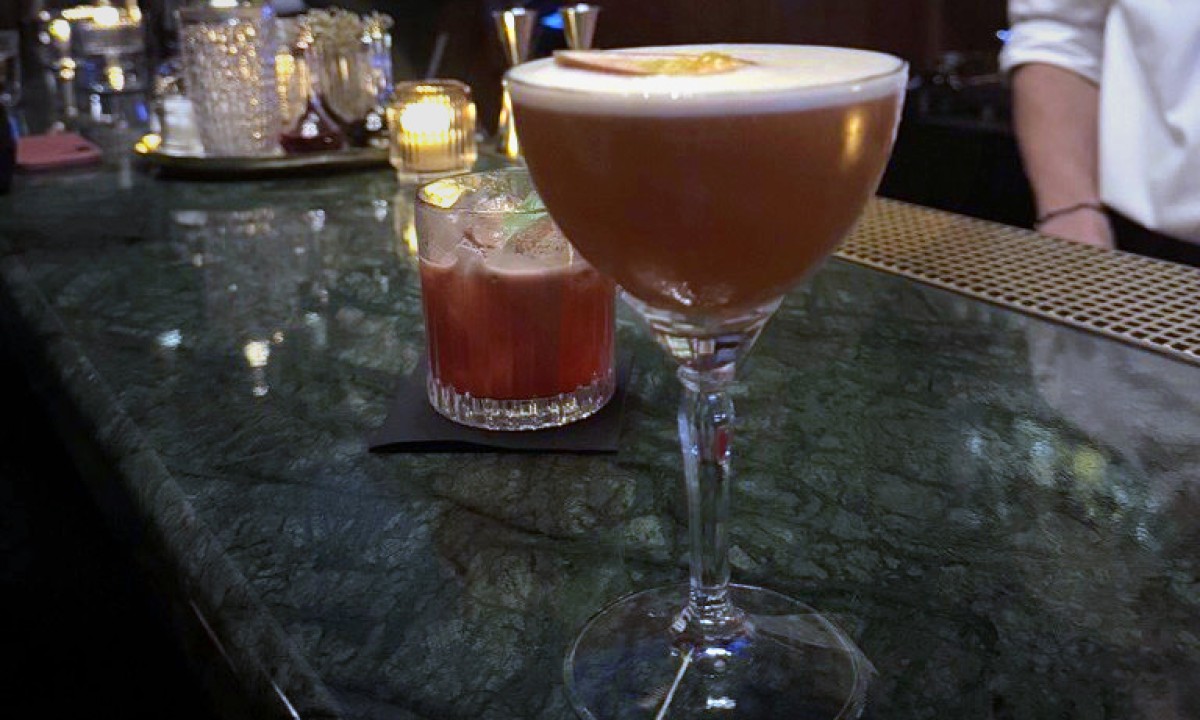 Zwei Cocktails auf einer dunklen Bar, einer in einem hohen Glas mit Schaum, der andere in einem kurzen Glas mit Eiswürfeln.