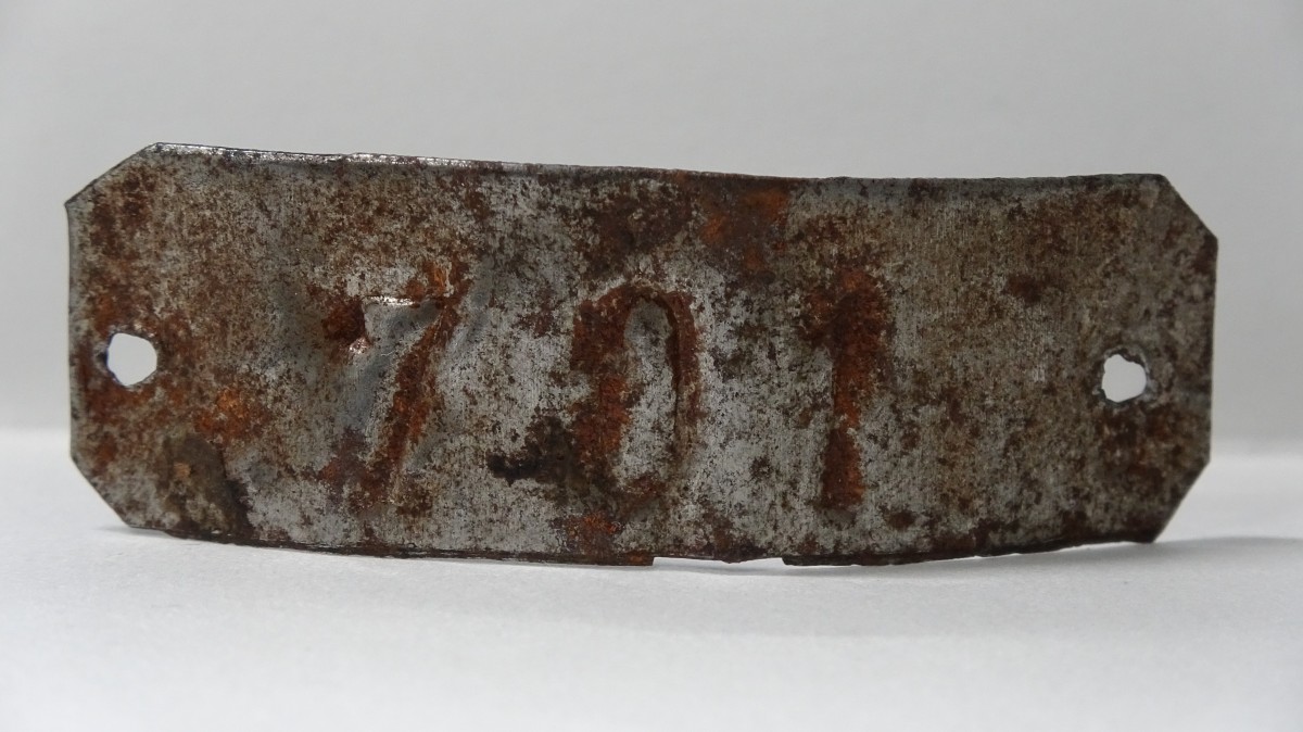 : Rusty metal plate with the number '701' embossed, featuring two holes on each side