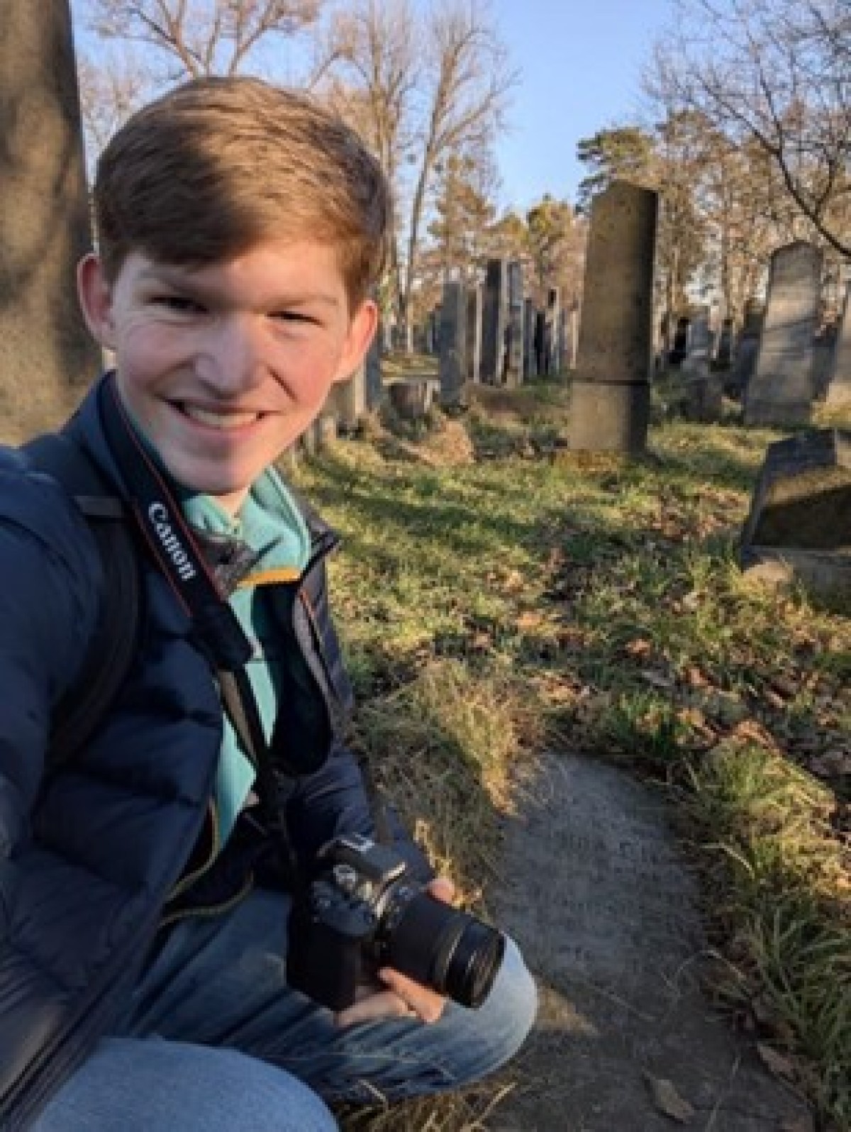 : Junger Mann mit Kamera kniet auf einem Friedhof mit alten Grabsteinen im Hintergrund