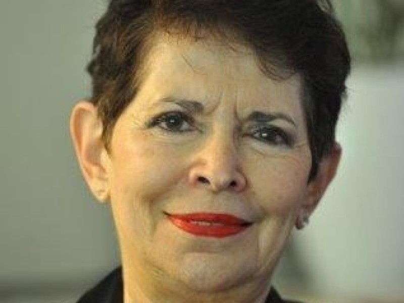 Portrait of an older woman with short dark hair wearing a black jacket and lace top, with a blurred background featuring flowers.