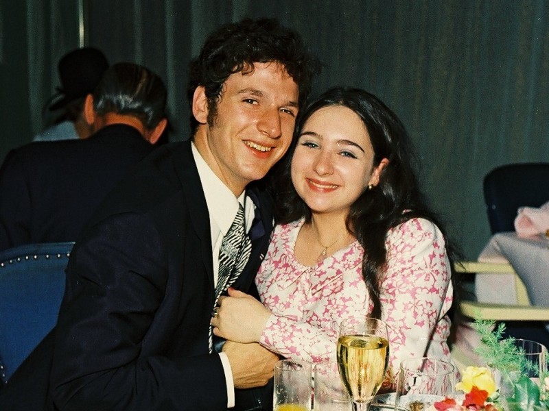 Junges Paar in festlicher Kleidung sitzt an einem Tisch mit Tellern, Getränken und Blumen und lächelt in die Kamera