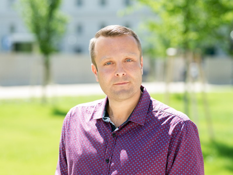 Mann mit kurzem, blondem Haar trägt ein lila Hemd mit kleinem Muster, steht im Freien vor unscharfem Hintergrund mit Bäumen und Gebäuden.