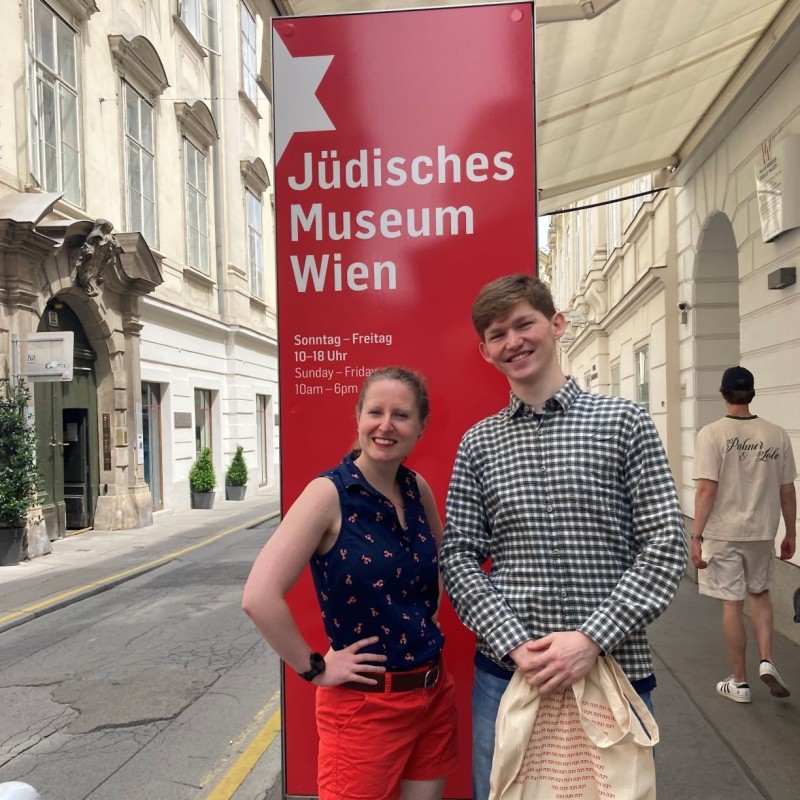 : Zwei lächelnde Personen stehen vor einem roten Schild mit der Aufschrift 'Jüdisches Museum Wien' und Öffnungszeiten in Deutsch und Englisch