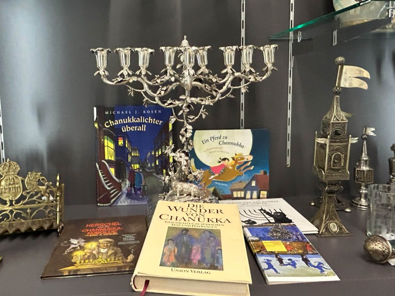 Mehrarmiger silberner Chanukkaleuchter, mehrere Chanukka-Bücher mit farbigen Covern, goldener Leuchter und dekorative Objekte in einer Vitrine.