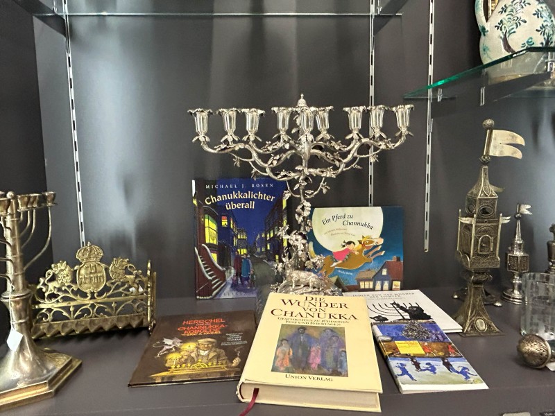 Mehrarmiger silberner Chanukkaleuchter, mehrere Chanukka-Bücher mit farbigen Covern, goldener Leuchter und dekorative Objekte in einer Vitrine.
