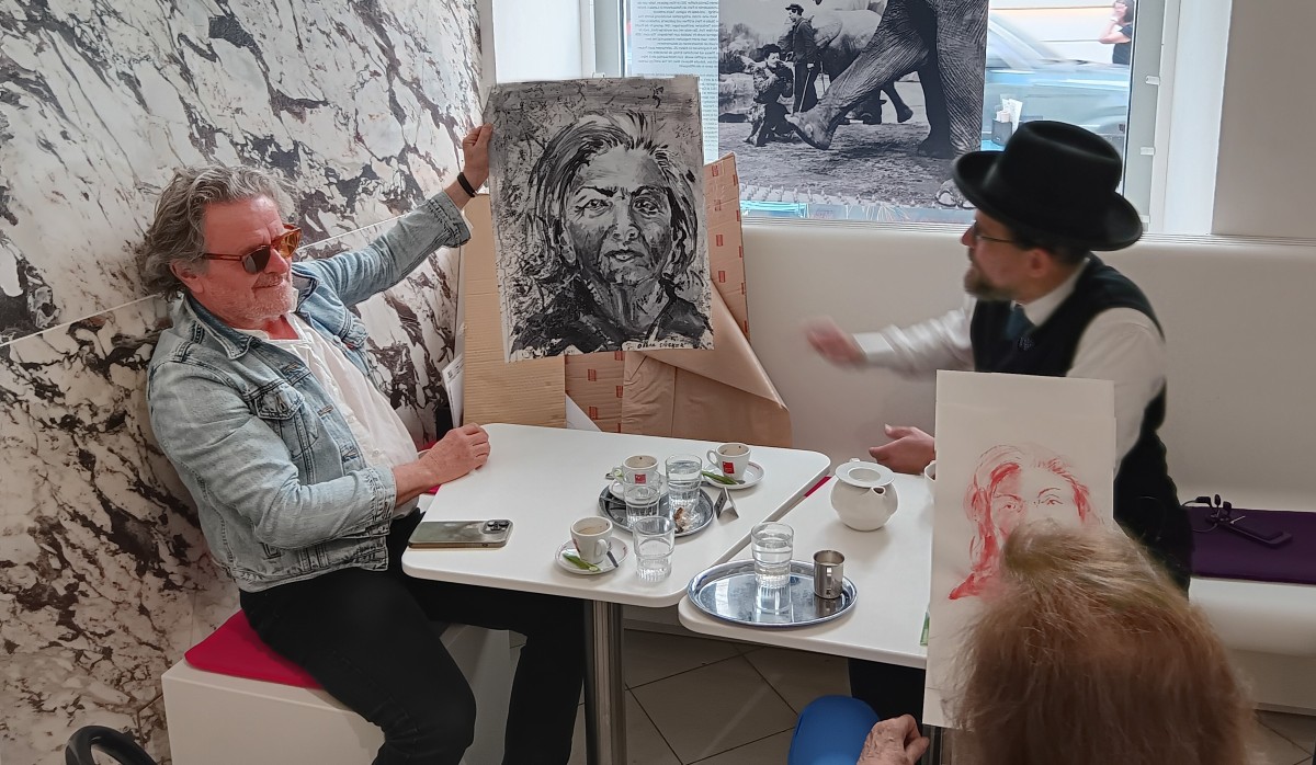 : Drei Personen sitzen an einem Tisch mit Kaffee und Wasser, zwei zeigen Zeichnungen von Gesichtern, eine Person trägt einen Hut und Weste.