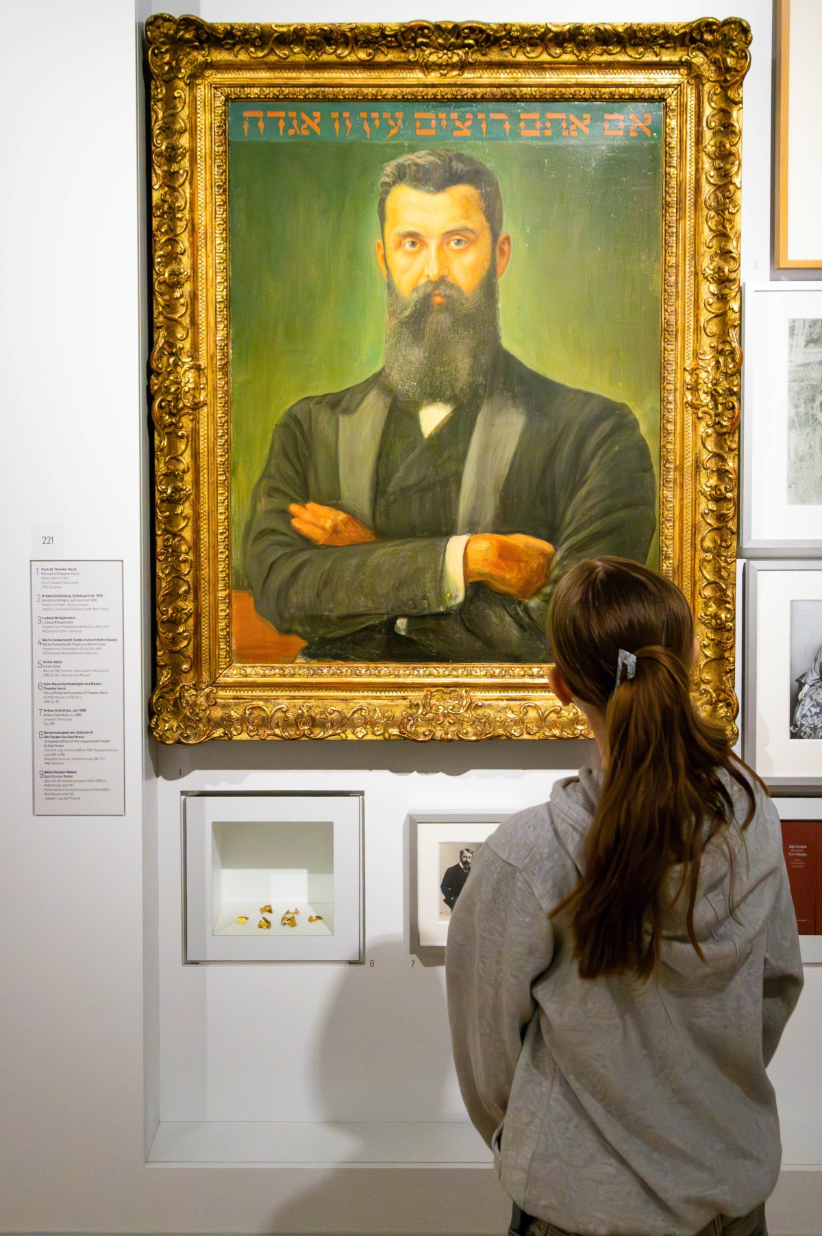 Porträt eines bärtigen Mannes in einem Museum: Eine Frau von hinten mit Zopf und grauem Pullover steht vor einem großen gemalten Porträt eines bärtigen Mannes mit verschränkten Armen in schwarzem Anzug, mit hebräischem Schriftzug oben.