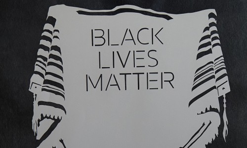 Ein weißer Tallit mit schwarzen Streifen, darauf steht 'BLACK LIVES MATTER'. Darunter hebräischer Text in schwarzer Schrift.