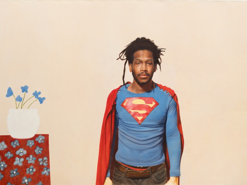 Mann mit Dreadlocks trägt ein blaues Superman-Shirt und einen roten Umhang, steht vor beige Hintergrund neben rotem Tisch mit weißer Vase und blauen Blumen