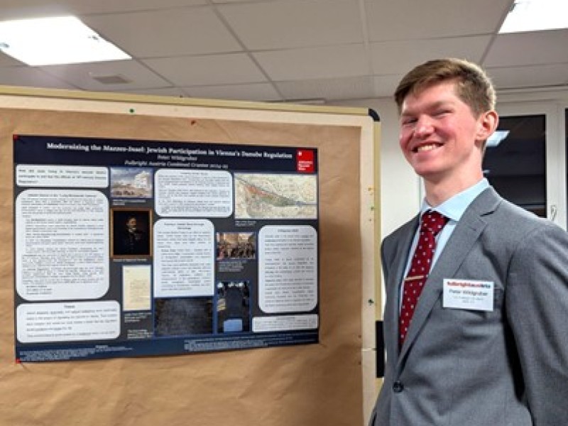 : Junger Mann in Anzug und roter Krawatte steht lächelnd neben einem wissenschaftlichen Poster über jüdische Beteiligung an der Wiener Donau-Regulierung