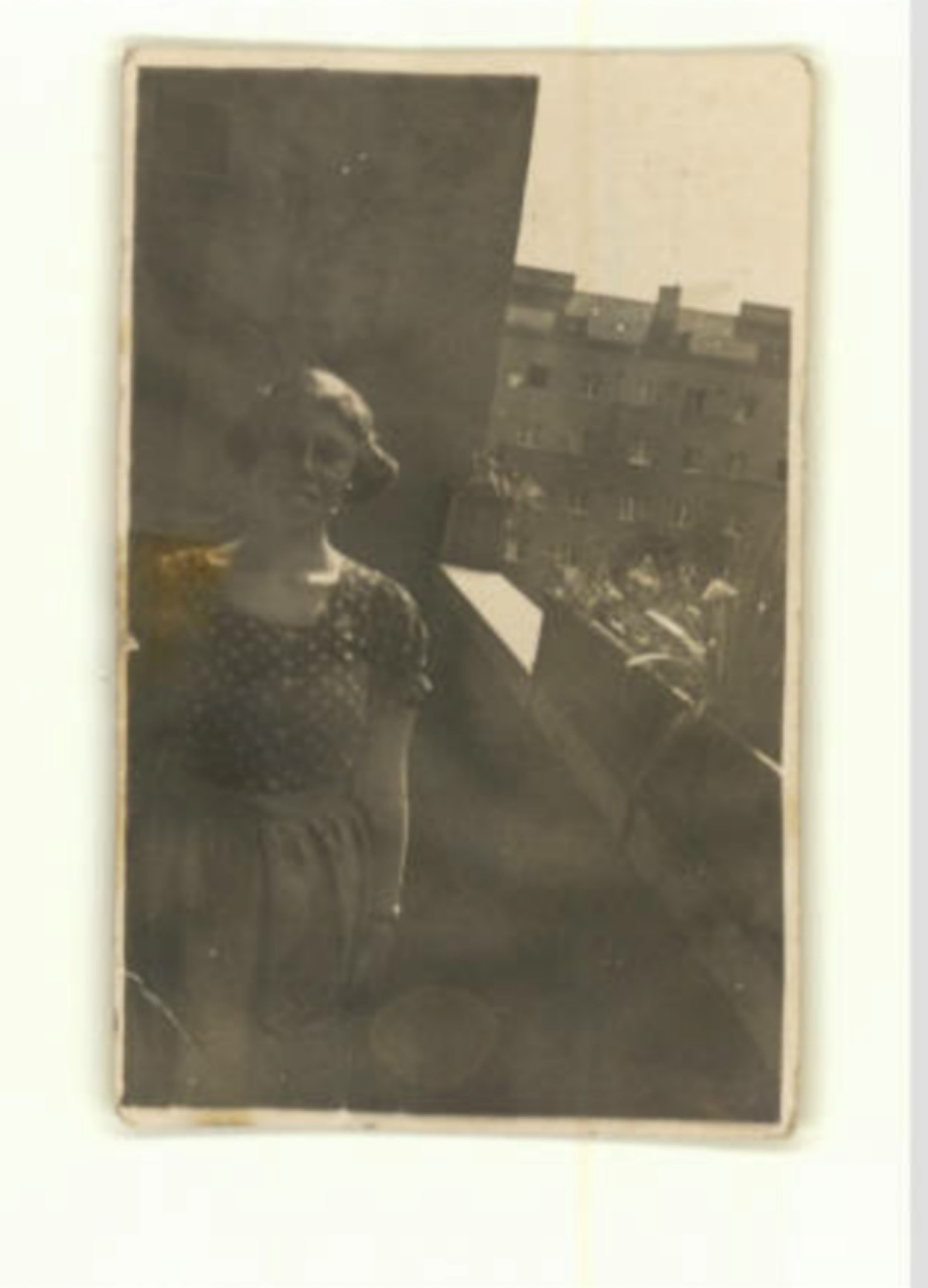 : Black and white photo of a woman standing on a balcony with apartment buildings in the background