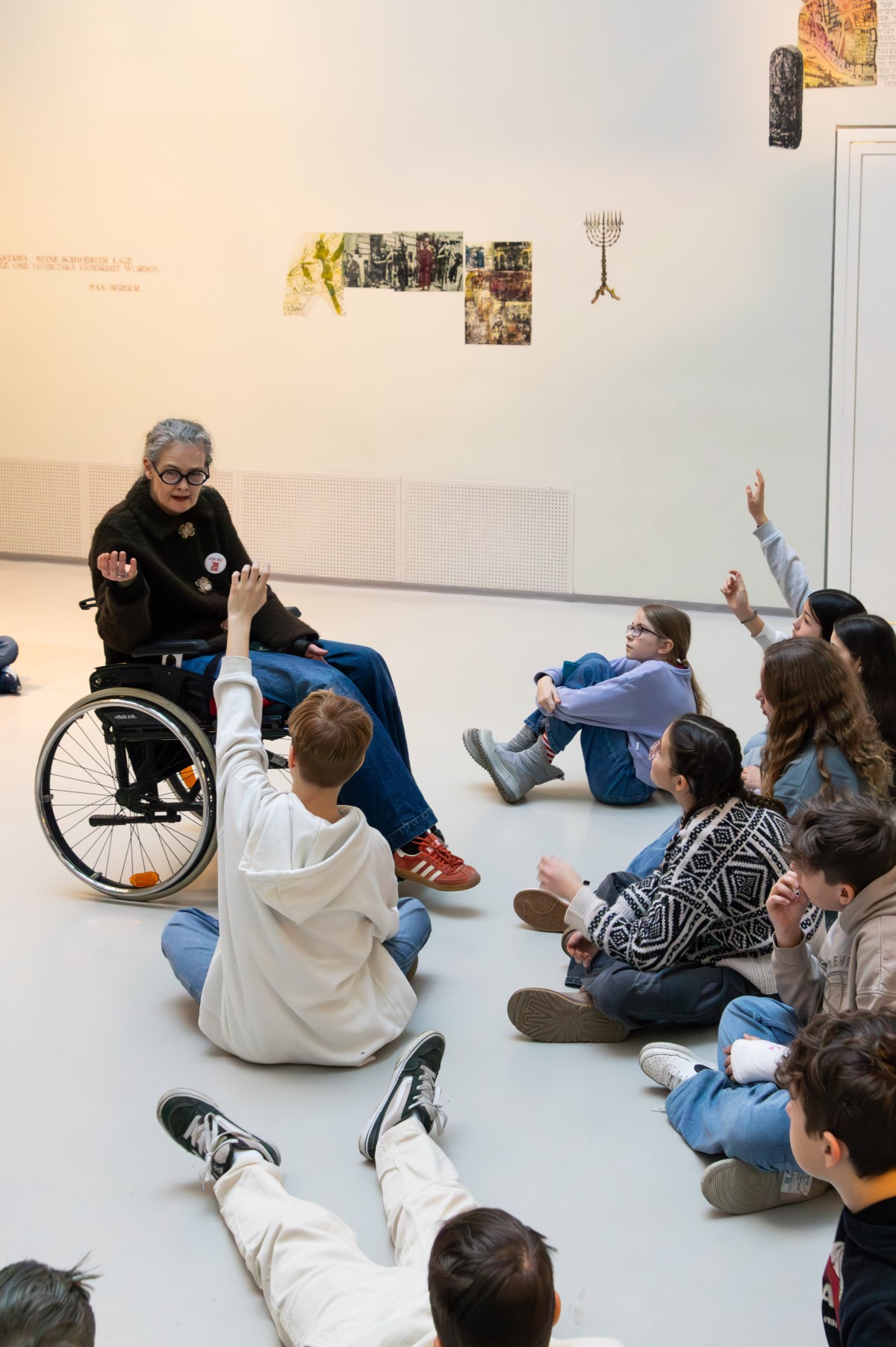 Führung im Museum mit Rollstuhlfahrerin und Kindern: Frau im Rollstuhl spricht mit einer Gruppe sitzender Kinder im Museum, zwei Kinder heben die Hand