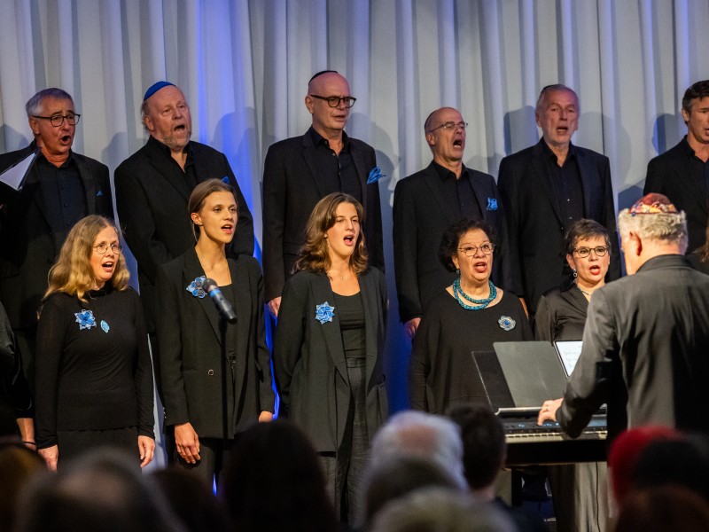 Chor mit elf Personen in schwarzer Kleidung singt unter Leitung eines Mannes am Klavier, einige Männer tragen Kippa