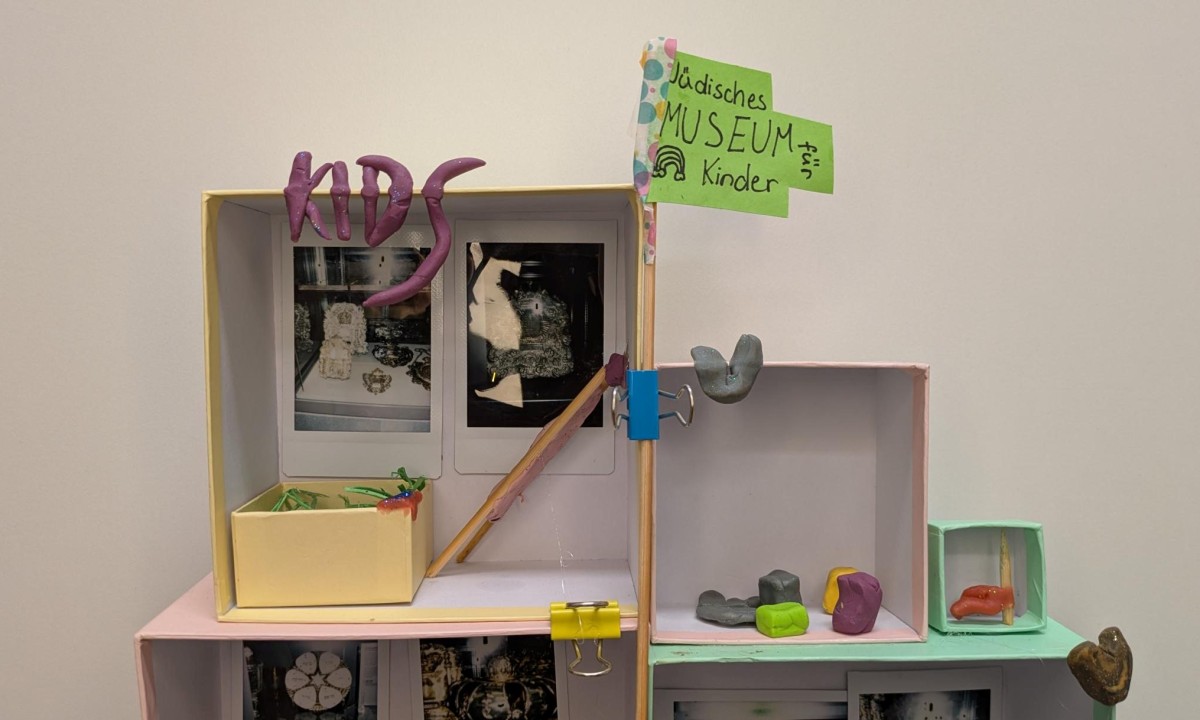 Kleines Diorama mit dem Wort "KIDS" aus lila Modelliermasse, zwei Fotos im Hintergrund und einem kleinen gelben Kasten mit grüner Deko im Vordergrund.