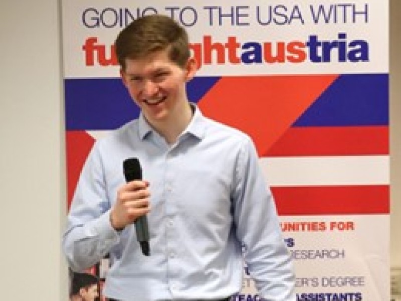 : Junger Mann in hellblauem Hemd hält Mikrofon und steht hinter einem Rednerpult mit Aufschrift 'fulbrightaustria Full of bright minds.' vor einem Banner mit Text 'GOING TO THE USA WITH fulbrightaustria'