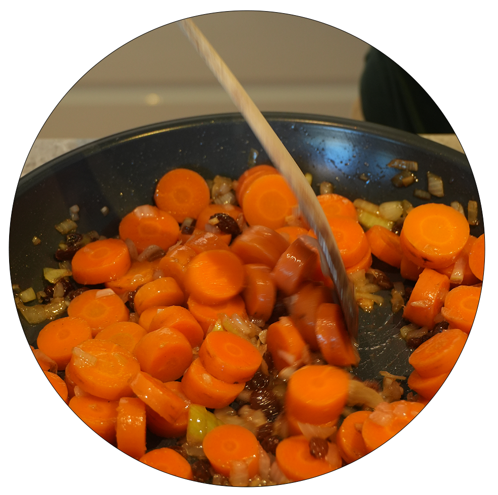 Eine Pfanne mit geschnittenen Karotten und Zwiebeln, die mit einem Holzlöffel umgerührt werden. Die Karotten sind orange und kreisförmig geschnitten.