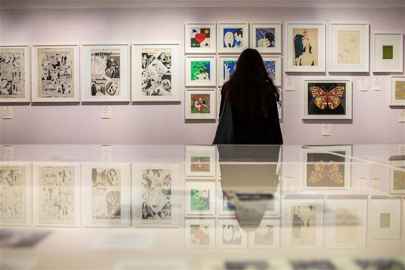 Person mit langen dunklen Haaren betrachtet eine Wand mit gerahmten Kunstwerken in einer Galerie, darunter ein Schmetterlingsbild